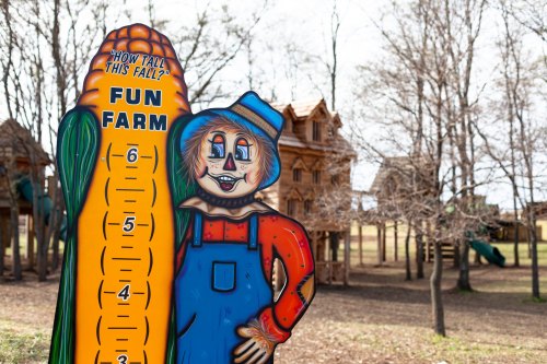 A colorful scarecrow sign next to a corn height chart at a fun farm, with wooden play structures in the background.
