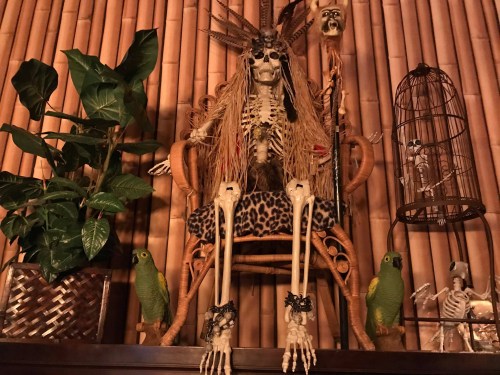 A decorated skeleton sits in a chair surrounded by plants, a birdcage, and two green parrot figures.