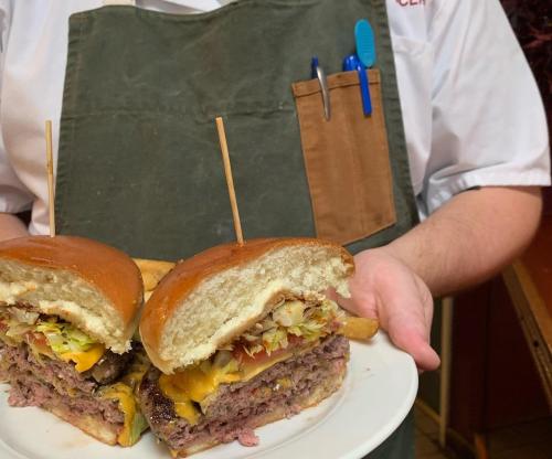 A chef holds a plate with two stacked burgers topped with cheese, lettuce, and condiments, alongside fries.