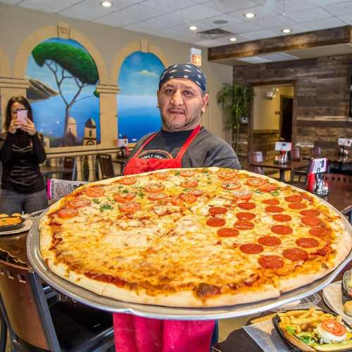 A chef in an apron holds a large pizza topped with cheese and pepperoni in a restaurant setting.
