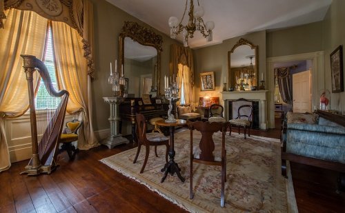 Elegant room with vintage decor, featuring a harp, mirrors, chandeliers, and ornate furniture on a patterned rug.