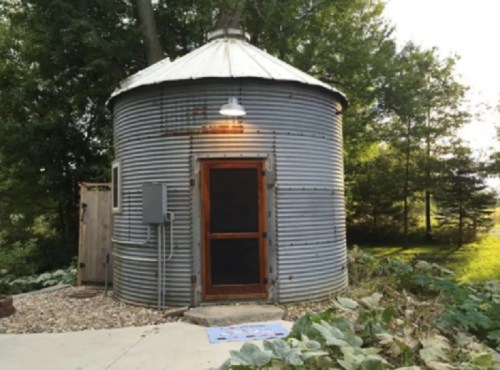A rustic, round metal grain silo converted into a small cabin, surrounded by greenery and a stone pathway.