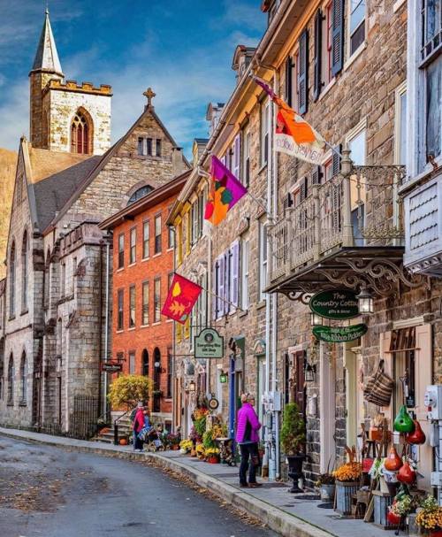 Charming street scene with historic buildings, colorful flags, and shops, set against a backdrop of a church steeple.