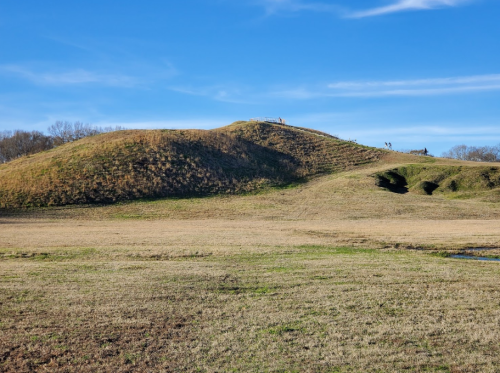 A grassy hill with a clear blue sky, featuring people at the top and a flat landscape in the foreground.