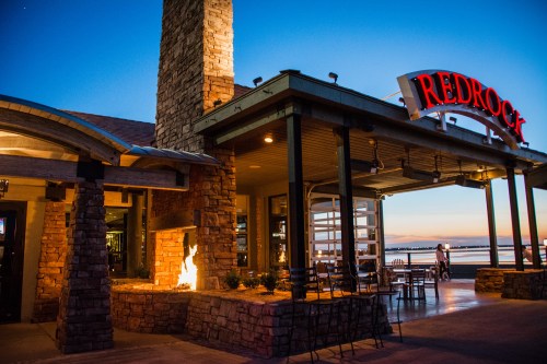 Exterior of Redrock restaurant at dusk, featuring stone architecture and a fire pit, with a scenic waterfront view.