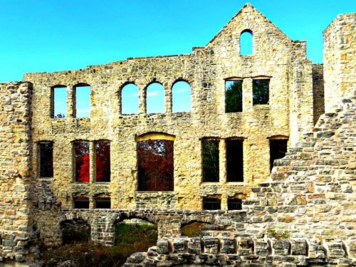 Ruins of a stone building with arched windows against a clear blue sky, surrounded by greenery and autumn foliage.