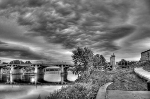 Black and white image of a river with a bridge, trees, and dramatic clouds reflecting on the water's surface.