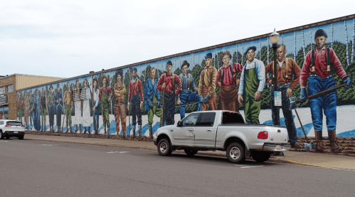 A mural depicting diverse figures in colorful clothing, standing along a wall, with parked vehicles in front.