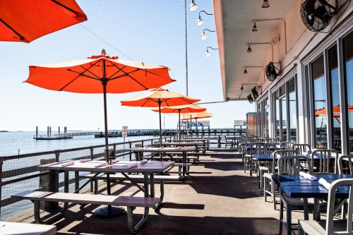 Outdoor dining area with orange umbrellas overlooking a calm waterfront and a pier in the distance.