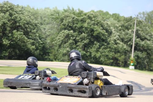 Two go-karts racing on a track, with drivers in helmets, surrounded by green trees and a clear blue sky.