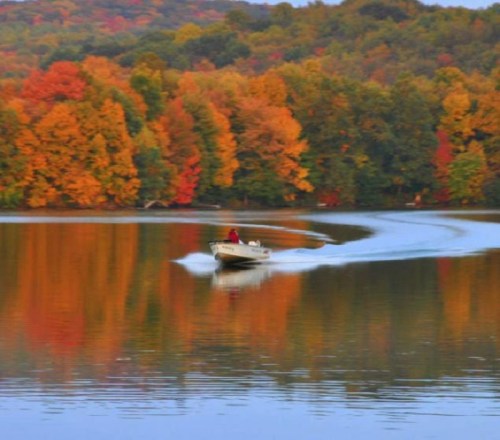 A boat glides across a calm lake, surrounded by vibrant autumn foliage reflecting in the water.
