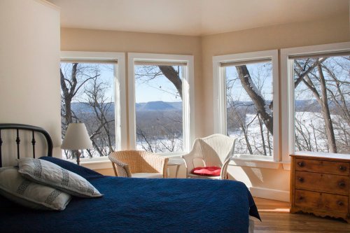 Cozy bedroom with a blue bedspread, large windows offering a view of trees and a river, and a wooden dresser.