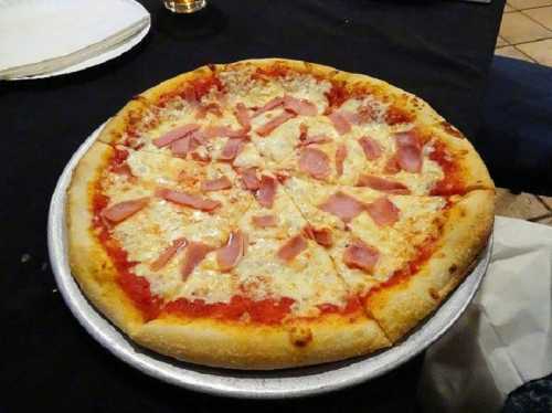 A round pizza topped with cheese and slices of ham on a silver tray, set on a black tablecloth.