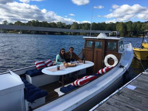 A couple enjoys a meal on a boat, surrounded by water and trees, with a sunny sky and clouds above.