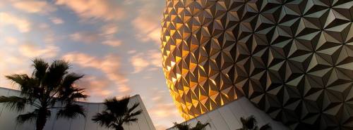A geometric structure with a golden hue against a colorful sky, surrounded by palm trees.