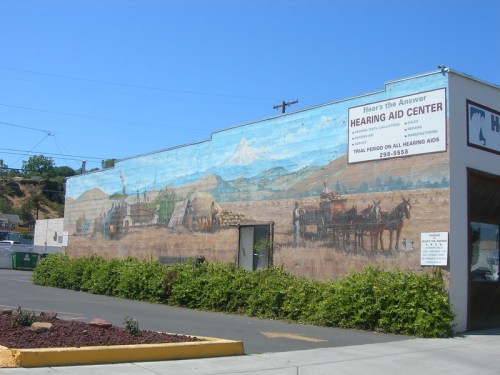 Mural on a building depicting a scenic landscape with mountains and horse-drawn wagons, alongside a hearing aid center sign.
