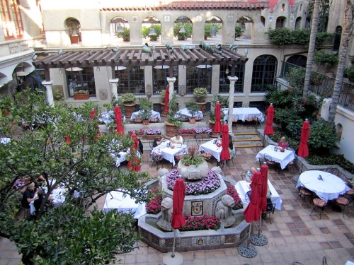 A charming outdoor dining area with tables, red umbrellas, and lush greenery surrounding a central fountain.