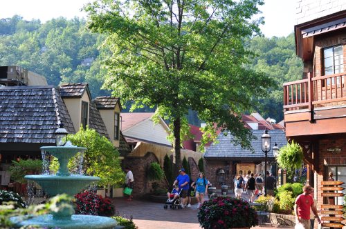 A charming village scene with shops, a fountain, and people strolling under a large tree in a lush, green setting.