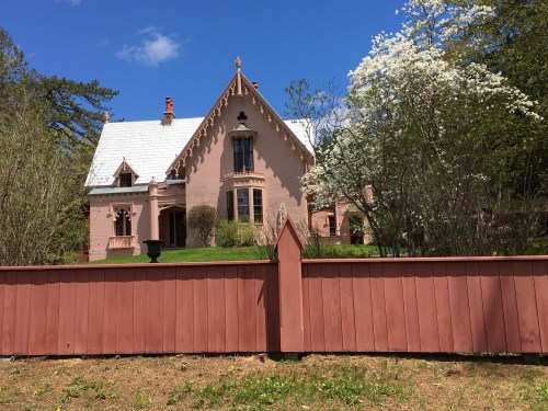 A pink Victorian-style house with a white roof, surrounded by trees and blooming flowers, behind a wooden fence.