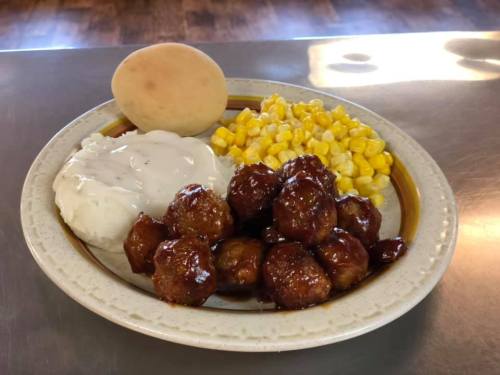 A plate with meatballs in sauce, mashed potatoes with gravy, corn, and a roll.