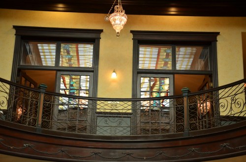 Interior view of a balcony with ornate railings and stained glass windows, illuminated by a hanging chandelier.