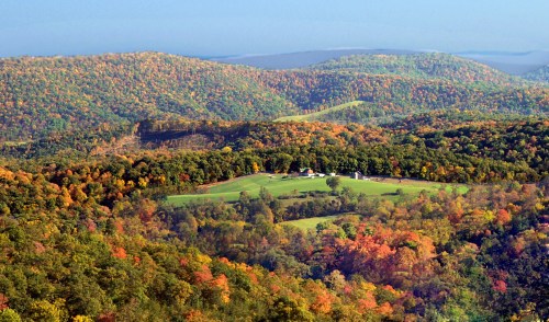 Aerial view of rolling hills covered in vibrant autumn foliage, with a green field and farmhouses nestled in the valley.