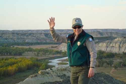 A park ranger waves from a scenic overlook, with rolling hills and a river in the background.