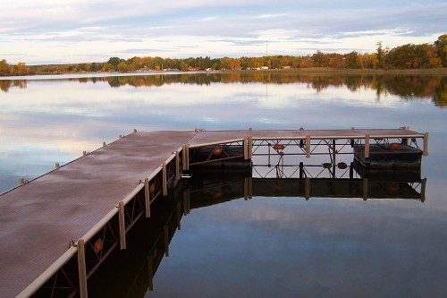 A serene lake scene with a wooden dock reflecting in calm waters, surrounded by autumn foliage and a cloudy sky.