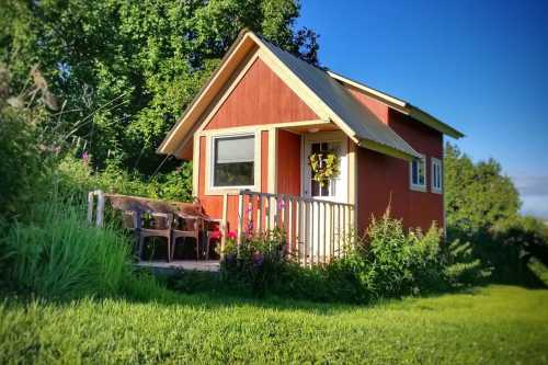 A small red cabin with a porch, surrounded by greenery and flowers under a clear blue sky.