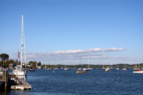 A serene harbor scene with sailboats and motorboats on calm water under a clear blue sky with scattered clouds.