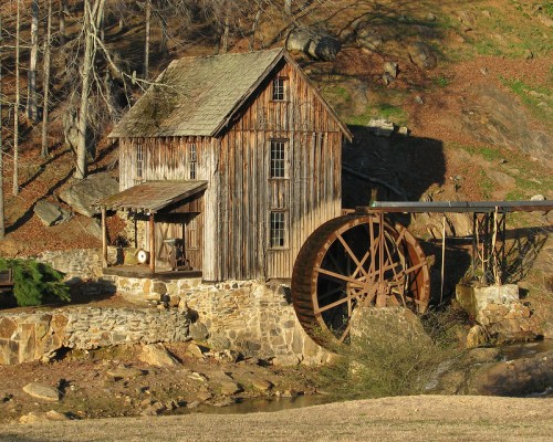 A rustic wooden mill with a large water wheel, set against a backdrop of trees and rocky terrain.