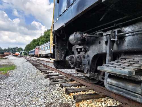 Close-up of a train's wheel and track, with vintage train cars in the background under a partly cloudy sky.