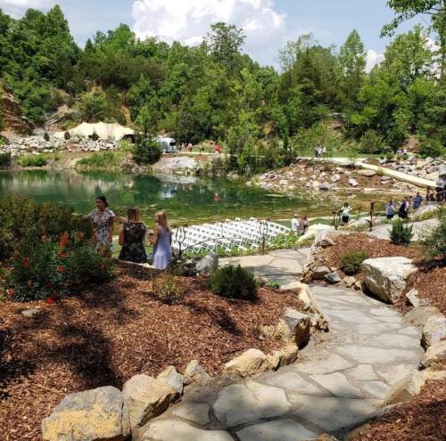 A scenic outdoor venue with a pond, surrounded by greenery and rocky paths, set up for an event with guests mingling.