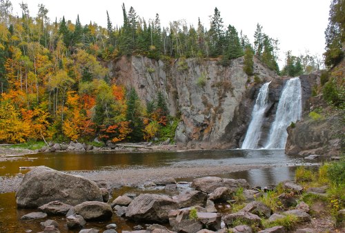 A serene waterfall cascades over rocky cliffs, surrounded by vibrant autumn foliage and a calm river.