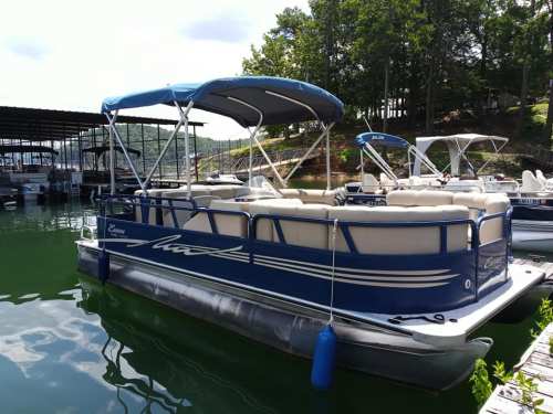A blue pontoon boat with a canopy is docked on green water, surrounded by trees and other boats in the background.