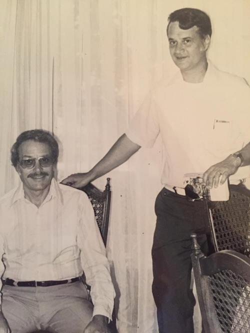 Two men pose for a photo, one seated with glasses and a mustache, the other standing beside him, both smiling.