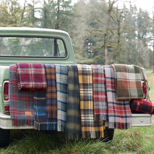A collection of colorful plaid blankets draped over the back of a vintage truck in a forested setting.