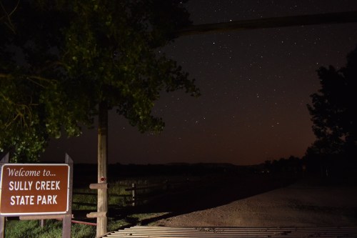 A sign reading "Welcome to Sully Creek State Park" under a starry night sky with a dirt road and trees in the foreground.