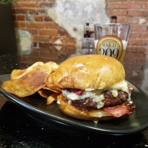 A gourmet burger with cheese and bacon, served with crispy potato chips and a glass of beer in the background.