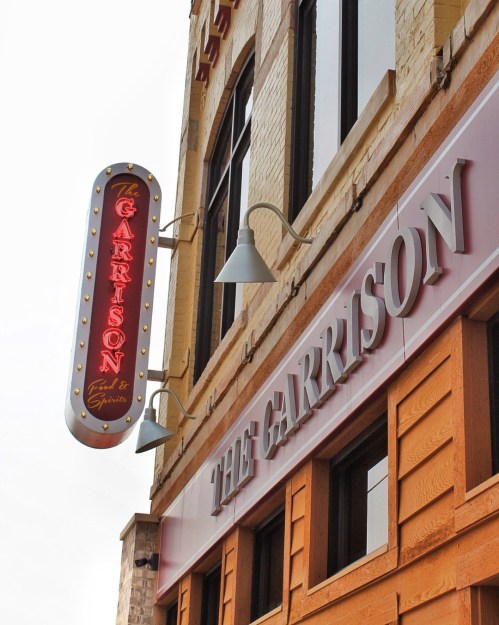 Sign for "The Garrison" with neon lights, mounted on a brick building.