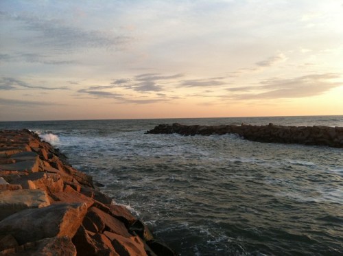 A rocky jetty extends into the ocean under a colorful sunset, with gentle waves lapping against the shore.