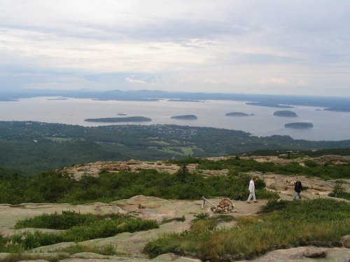 A scenic view from a mountain overlooking a lake with islands, featuring two hikers on a rocky path.