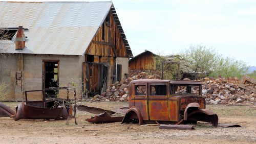 An old, rusted car sits in front of a weathered barn, surrounded by debris and overgrown vegetation.