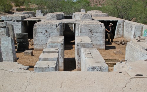 Ruins of a concrete structure in a desert landscape, with a person exploring the site among the weathered walls.