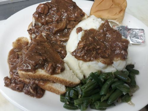 A plate of mashed potatoes with gravy, sliced bread topped with meat, green beans, and a bun on the side.