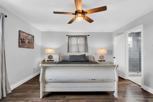 Cozy bedroom with a white sleigh bed, ceiling fan, lamps, and a bathroom door in the background. Neutral color scheme.