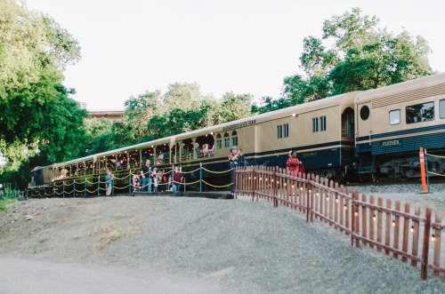 A vintage train parked beside a gravel path, with people gathered on the platform and trees in the background.