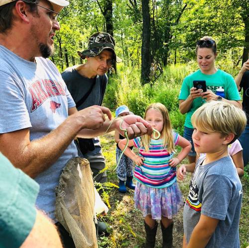 A group of people watches as a man holds a snake, engaging children in a nature setting.