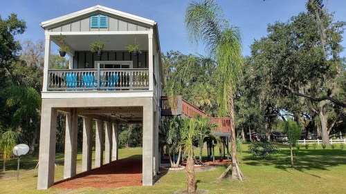 A modern elevated house with a porch, surrounded by palm trees and greenery, set in a spacious outdoor area.