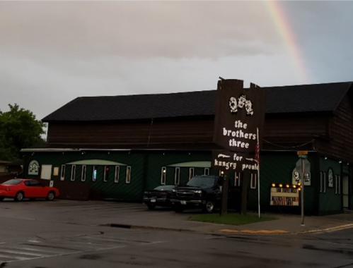 A rustic restaurant named "The Brothers Three" with a rainbow in the background and parked cars in front.